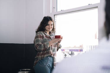 Lächelnde Geschäftsfrau mit Kaffeetasse, die in der Nähe eines Fensters im Büro steht - AMWF01121