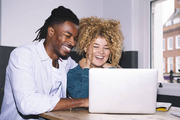 Happy businessman sitting with colleague looking at laptop in office - AMWF01127