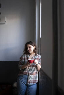 Lächelnde Geschäftsfrau mit Kaffeetasse, die am Fenster im Büro steht - AMWF01132