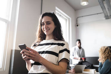 Happy businesswoman holding smart phone with colleagues in background - AMWF01137