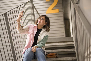 Glückliche Frau nimmt Selfie durch Smartphone sitzen auf Treppen - JJF00272