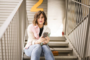 Selbstbewusste Frau mit Smartphone auf der Treppe sitzend - JJF00273