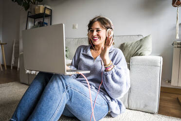 Happy woman waving through laptop sitting on floor at home - JJF00280