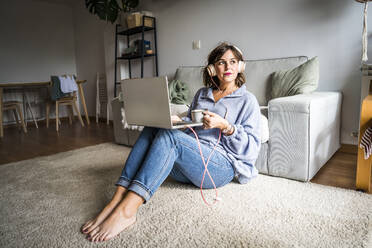 Nachdenkliche Frau sitzt mit Laptop und Kaffeetasse zu Hause - JJF00281