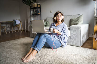 Thoughtful woman sitting with laptop and coffee cup at home - JJF00281