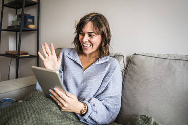 Glückliche Frau bei einem Videogespräch, die durch einen Tablet-PC auf dem Sofa zu Hause winkt - JJF00289