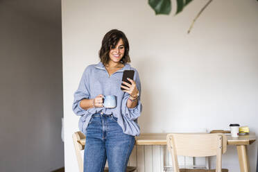 Happy woman with coffee cup using smart phone at home - JJF00297