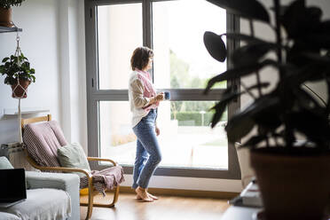 Frau mit Kaffeetasse am Fenster stehend zu Hause - JJF00308