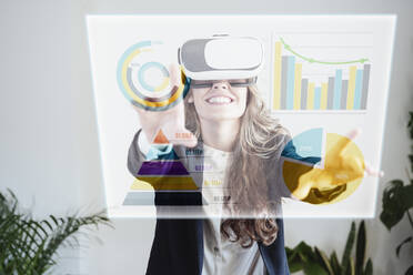 Happy businesswoman wearing VR goggles and examining graphs and pie charts - EBBF08092