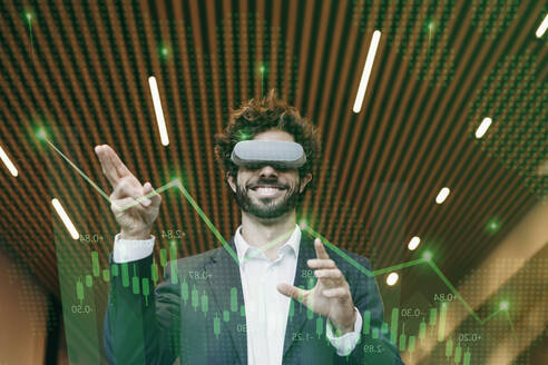 Glücklicher Geschäftsmann, der einen Virtual-Reality-Simulator trägt und im Büro gestikuliert - EBBF08099