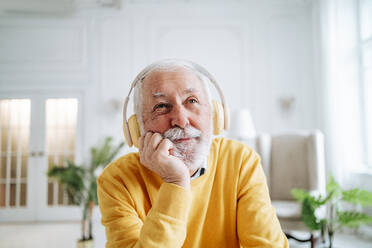 Nachdenklicher älterer Mann, der zu Hause über Kopfhörer Musik hört - MDOF00660