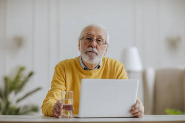 Senior freelancer with laptop and glass of water sitting at desk - MDOF00666