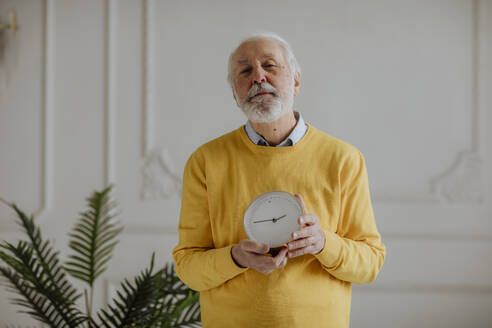 Senior man with clock standing in front of wall at home - MDOF00673