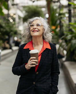 Nachdenklich lächelnde reife Geschäftsfrau mit Kaffeetasse im Garten stehend - JCCMF09512