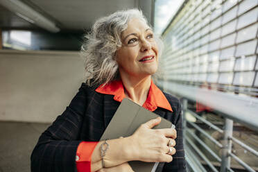 Thoughtful mature businesswoman with tablet PC looking through window - JCCMF09528