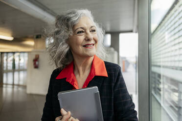 Happy mature businesswoman with tablet PC looking through window - JCCMF09541