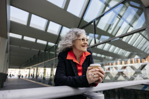 Happy mature businesswoman with disposable coffee cup leaning on railing - JCCMF09546