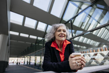 Geschäftsfrau mit grauem Haar hält Einweg-Kaffeebecher auf Geländer - JCCMF09548
