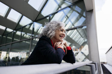Happy businesswoman with gray hair leaning on railing at station - JCCMF09550