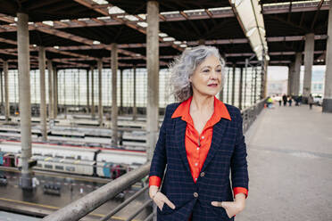 Mature businesswoman with hands in pockets walking at railroad station - JCCMF09556