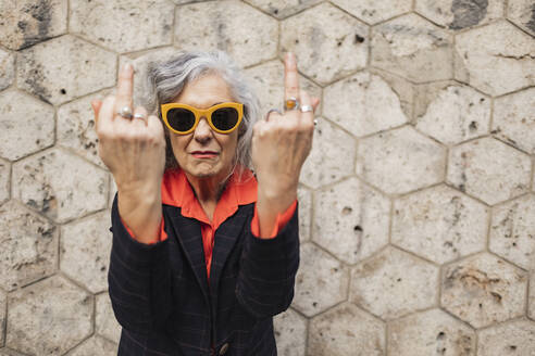 Mature businesswoman with gray hair showing obscene gesture in front of wall - JCCMF09566