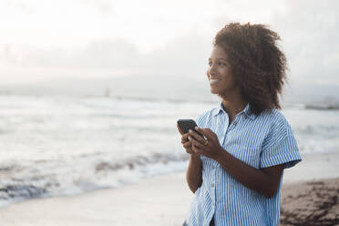 Glückliche Frau mit Smartphone am Strand stehend - JOSEF17249