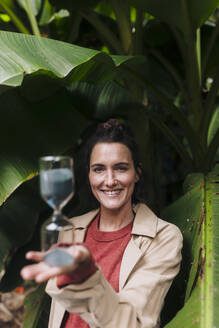 Happy woman with hourglass standing in front of banana tree - JOSEF17400