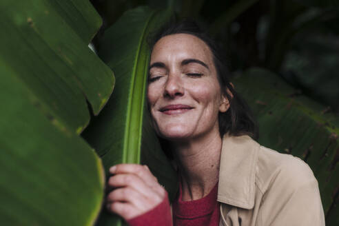 Lächelnde Frau mit geschlossenen Augen, die ein Bananenblatt umarmt - JOSEF17407
