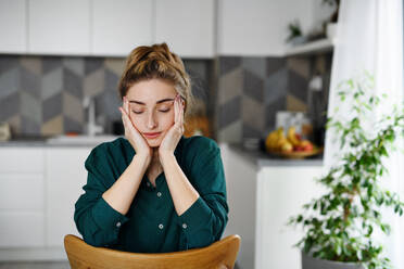 Ein Porträt einer müden jungen Studentin, die mit geschlossenen Augen in einer Küche sitzt und sich entspannt. - HPIF06145