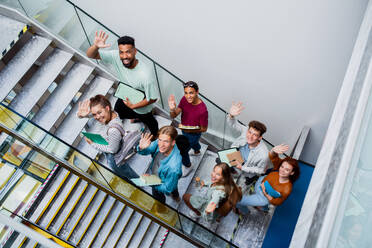 Blick von oben auf Studenten, die die Treppe in einem Gebäude hinaufgehen, in die Kamera schauen und winken. - HPIF06189