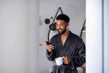 A young man with coffee and bathrobe indoors in bathroom at home, morning routine concept. - HPIF06250