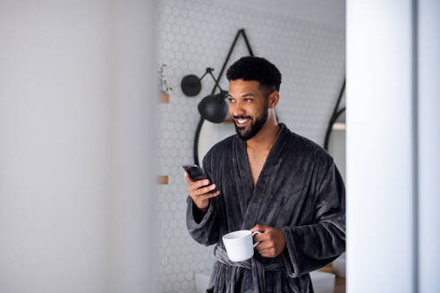 Ein junger Mann mit Kaffee und Bademantel drinnen im Badezimmer zu Hause, Morgenroutine Konzept. - HPIF06250