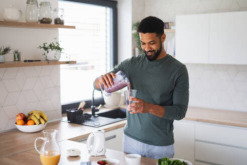 A young man preparing healthy breakfast indoors at home, pouring milk shake into glass. - HPIF06279