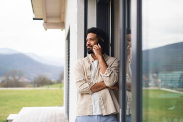 Porträt eines jungen Mannes, der mit seinem Smartphone im Freien auf der Terrasse seines Hauses telefoniert. - HPIF06286