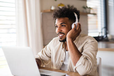 Portrait of young man student with laptop and headphones indoors at home, studying. - HPIF06298