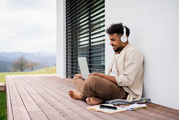 A young man student with laptop and headphones sitting on patio outdoors at home, studying. - HPIF06300