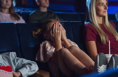 Ein besorgtes kleines Mädchen mit einer Mutter, die sich im Kino die Augen zuhält, während sie einen Film sieht. - HPIF06310