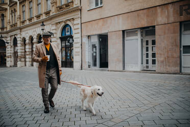 An elegant senior man with take away coffee walking his dog outdoors in city in winter. - HPIF06341