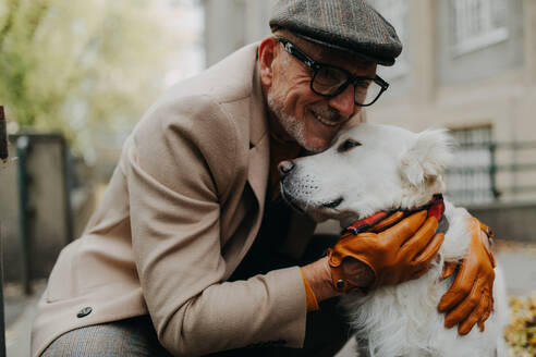 A happy senior man sitting on bench and embracing his dog outdoors in park in city. - HPIF06346