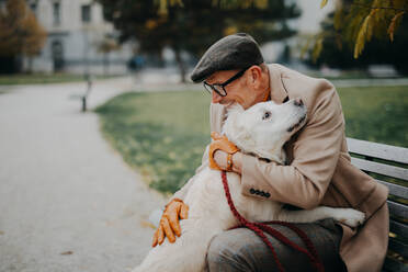 A happy senior man sitting on bench and embracing his dog outdoors in park in city. - HPIF06351