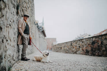 Ein älterer Mann schaut in die Kamera und lehnt sich an die Wand, während er mit seinem Hund in der Stadt spazieren geht. - HPIF06367