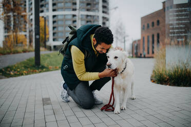 A happy young man squatting and stroking his dog during walk outdoors in city. - HPIF06372