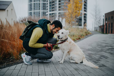 A happy young man squatting and embracing his dog during walk outdoors in city. - HPIF06373