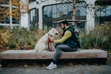 Seitenansicht eines jungen Mannes, der auf einer Bank sitzt und seinen Hund draußen in der Stadt trainiert. - HPIF06374