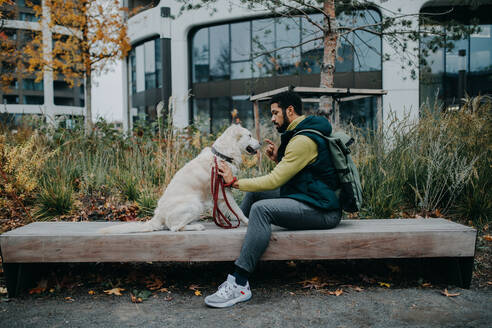 Seitenansicht eines jungen Mannes, der auf einer Bank sitzt und seinen Hund draußen in der Stadt trainiert. - HPIF06374