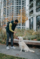 A side view of happy young man training his dog outdoors in city. - HPIF06376