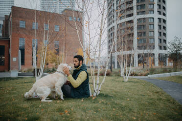 A happy young man sitting on grass with his dog outdoors in town. - HPIF06379