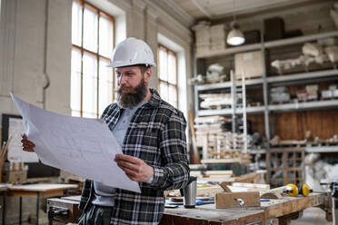 A mature male carpenter looking at blueprints plans in carpentery workshop. Small business concept. - HPIF06396