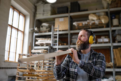 Reifer männlicher Tischler mit Gehörschutz, der ein Holzbrett trägt, drinnen in einer Tischlerwerkstatt, Konzept eines kleinen Unternehmens. - HPIF06402