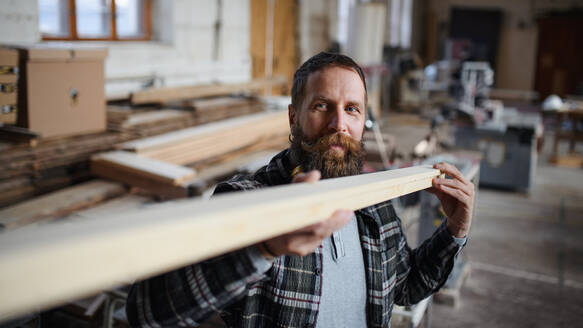 Ein reifer männlicher Schreiner trägt ein Holzbrett in einer Schreinerwerkstatt, ein kleines Geschäftskonzept. - HPIF06404